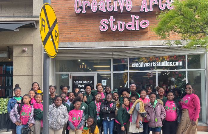 A group of girls and teachers from Mann pose for a picture in front of creative arts studion 
