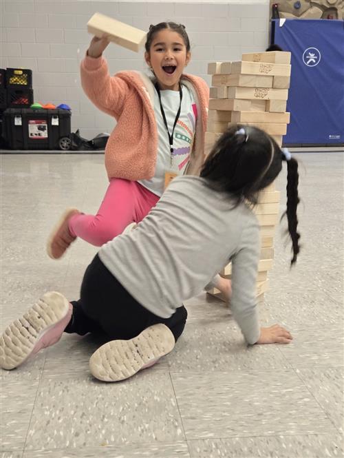 2 girls playing a large wooden stacking game on a gym floor
