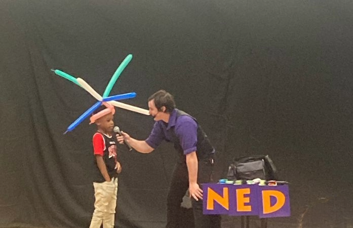  1st grade student on stage with Ned performer holding a microphone to the student