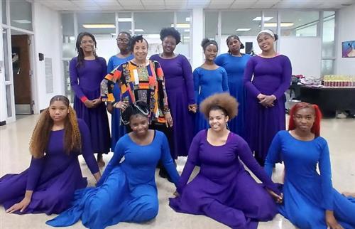  Dance teacher standing next to 6 students standing in back and 4 sitting on floor in blue 