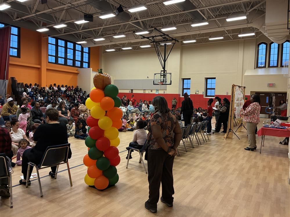  students sitting on gym floor, green brown orange balloons and teacher standing watch