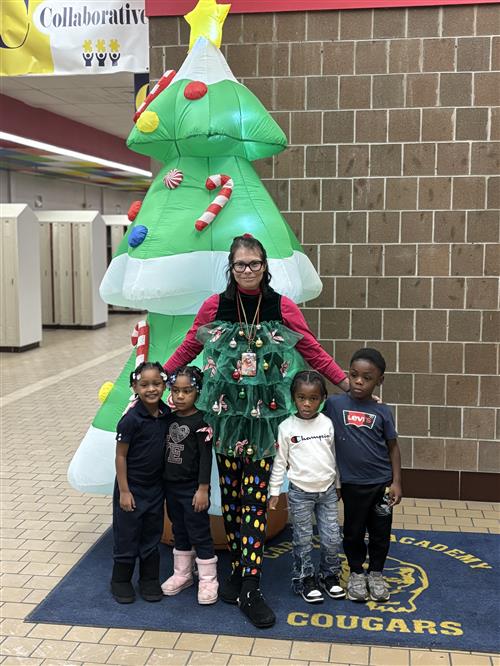 Four young students pose in front of a Tree prop while an adult dressed decoratively smiles in between them. 