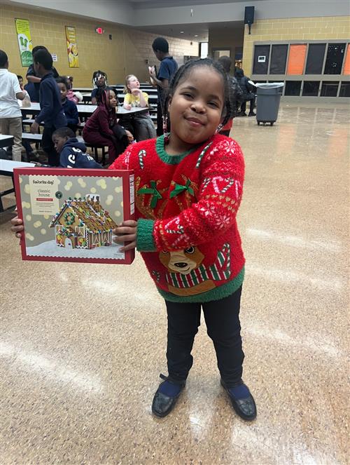 a young girl dressed in holiday gear posed with a gingerbread house kit. 