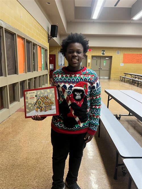 a student dressed in holiday gear posed with a gingerbread house kit. 