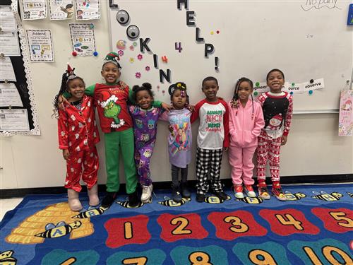 7 students posed in pajamas, smiling inside of a classroom standing on a numbered rug.