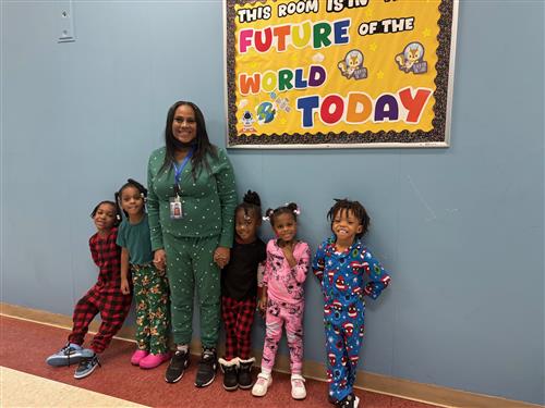 5 students along with an adult wearing pajamas and smiling in front of a blue wall with a bulletin board behind them. 