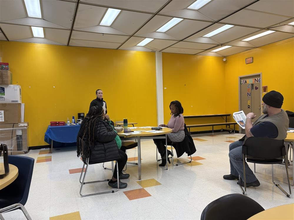  3 adults sitting at round tables listening to an adult standing.