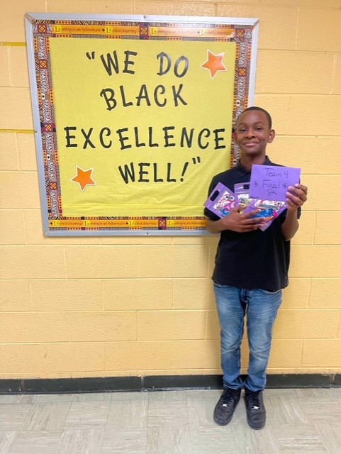  boy standing and smiling, holding prizes in front of a bulletin board