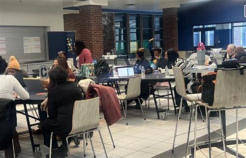 A group of people sitting in the lunchroom. They are listening to the female speaking standing.