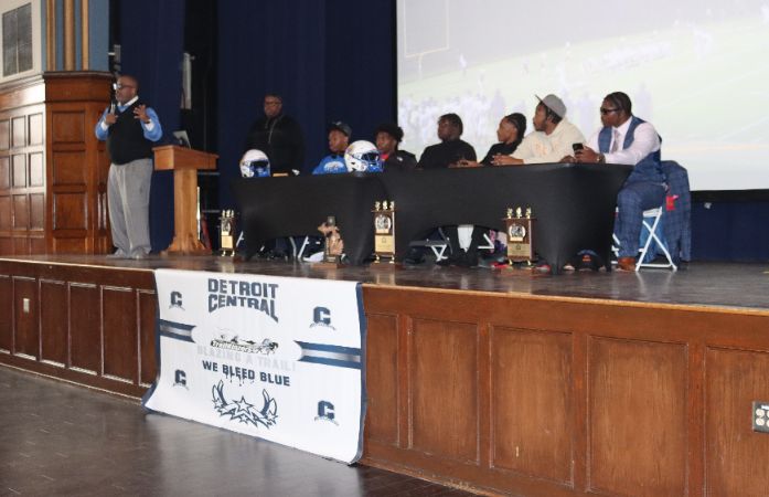  A man speaking, a man standing and 6 students are sitting at a table.