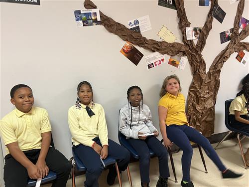 4 students sitting in chairs in front of poem-tree and smiling at camera