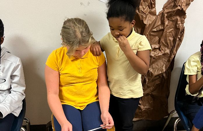 young student standing next to 4th grade student who is sitting and listening to her read her poem