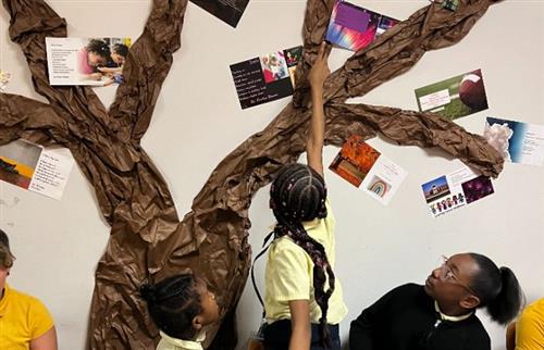 student reaching up and pointing at poem on poem-tree while two other students look up at her