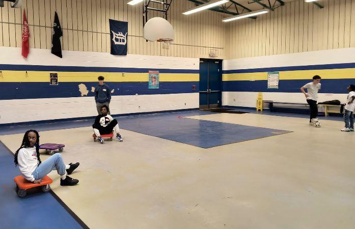 students in gym sitting on scooters and talking to high school students