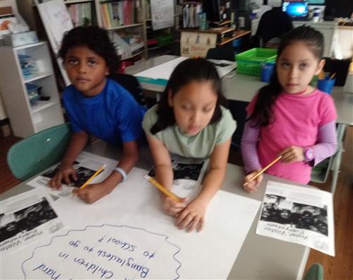 three 2nd grade students writing responses on chart paper 